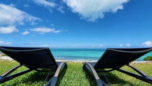 Two lounge chairs facing a clear blue sea with a grassy foreground and a stone wall.