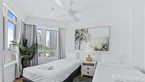 Een moderne hotelkamer met een groot bed, een klein bed, een plafondventilator, een nachtkastje met een lamp, uitzicht op de stad door grote ramen en een tropisch schilderij aan de muur.