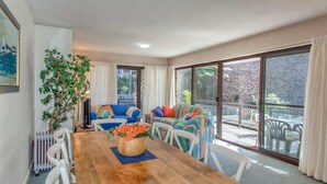 A dining area with a wooden table, white chairs, and colorful sofas. There is a potted plant on the table and a view of the outside through large glass doors. - Bay Parklands, 62 2 Gowrie Avenue - Wifi, Pool, Tennis, Water Views & Aircon (Nelson Bay)