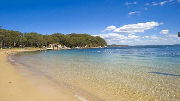 A beach with clear shallow waters, a sandy shore, and a building with a red roof near the water's edge. - Bay Parklands, 62 2 Gowrie Avenue - Wifi, Pool, Tennis, Water Views & Aircon (Nelson Bay)