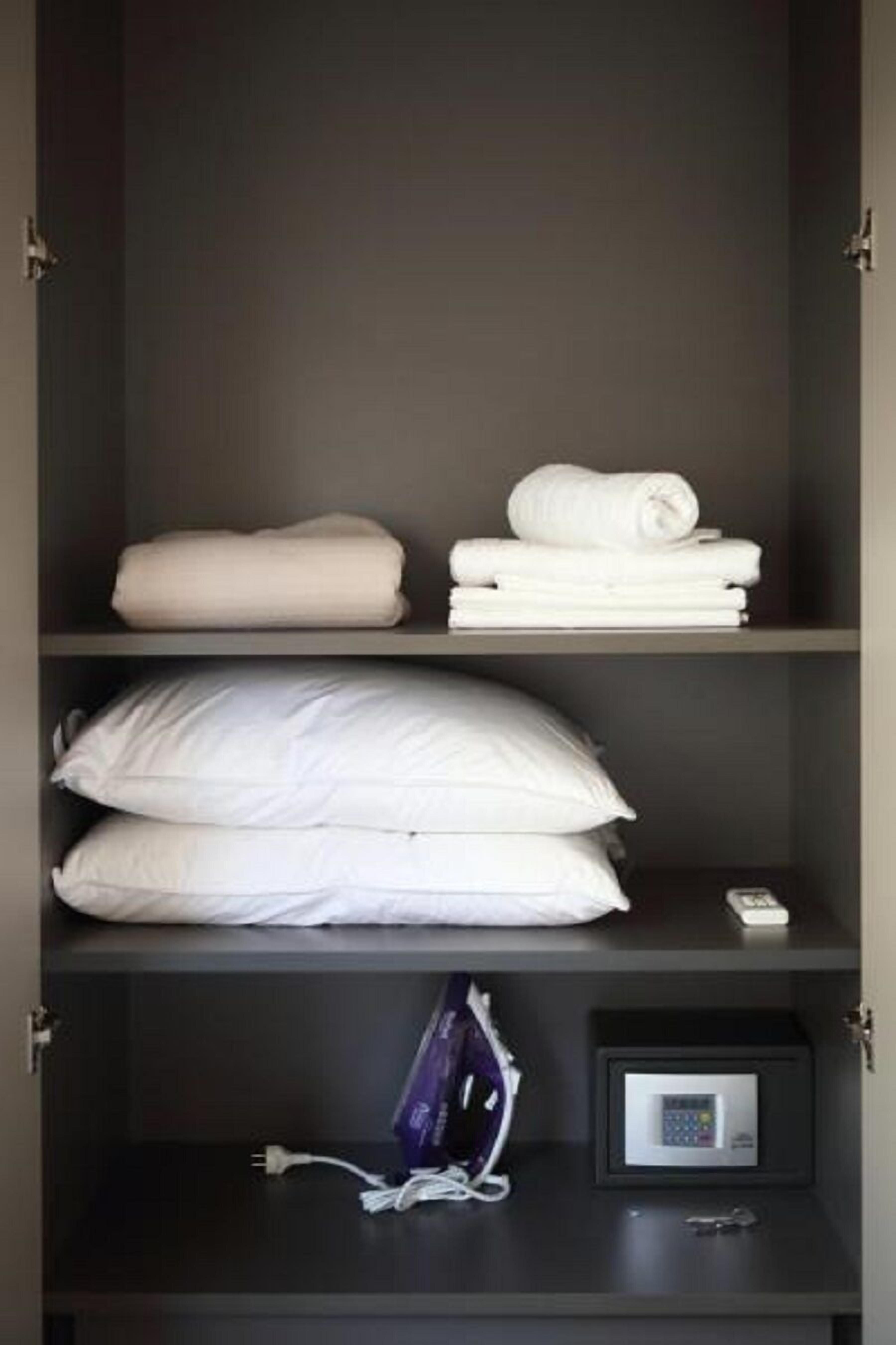 A hotel room with a dark-colored wardrobe containing neatly folded towels, a stack of pillows, a remote control, and a small safe with a key.
