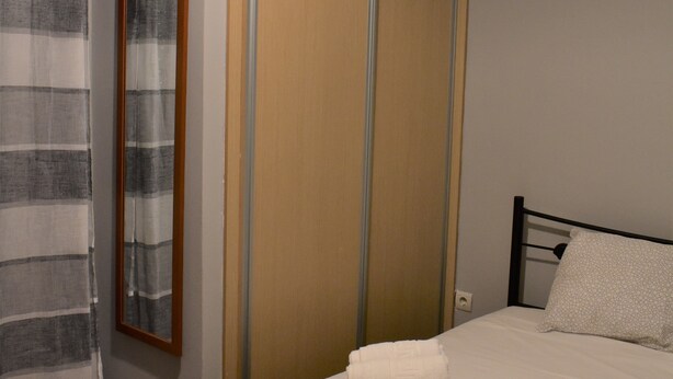 A neatly made bed with white linens and rolled towels, a wooden wardrobe, and a striped curtain. - Small & Cozy Flat near Hospitals (Zografos)