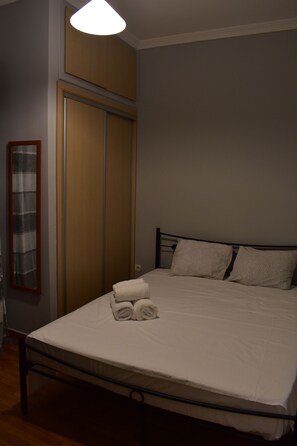 A neatly made bed with pillows and a rolled towel, a wooden wardrobe, and a mirror on the wall. - Small & Cozy Flat near Hospitals (Zografos)