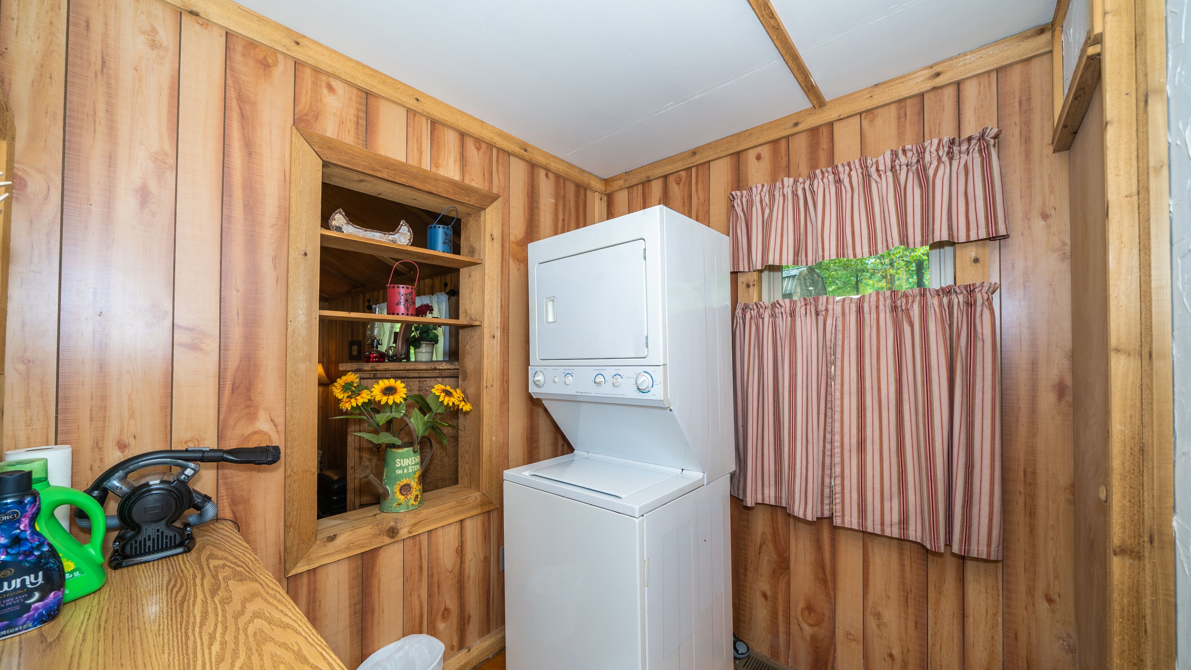 Una lavandería con paredes de madera, una lavadora, una ventana con repisa y una cortina.