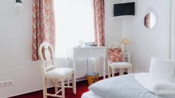 A hotel room with a bed, a desk with a chair, a small table with a lamp, a TV mounted on the wall, and a window with curtains.