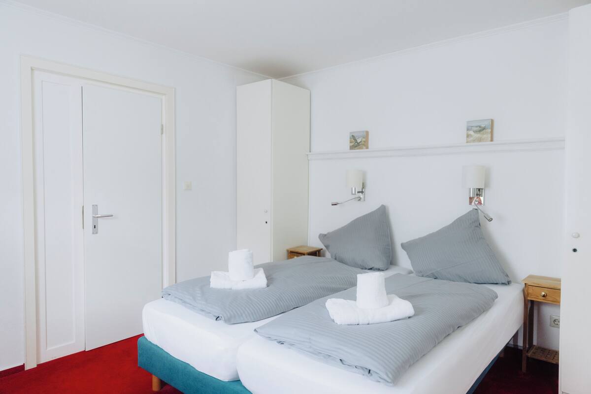 Una cama doble con dos almohadas y dos toallas, una mesita de noche, una lámpara de pared y una puerta.