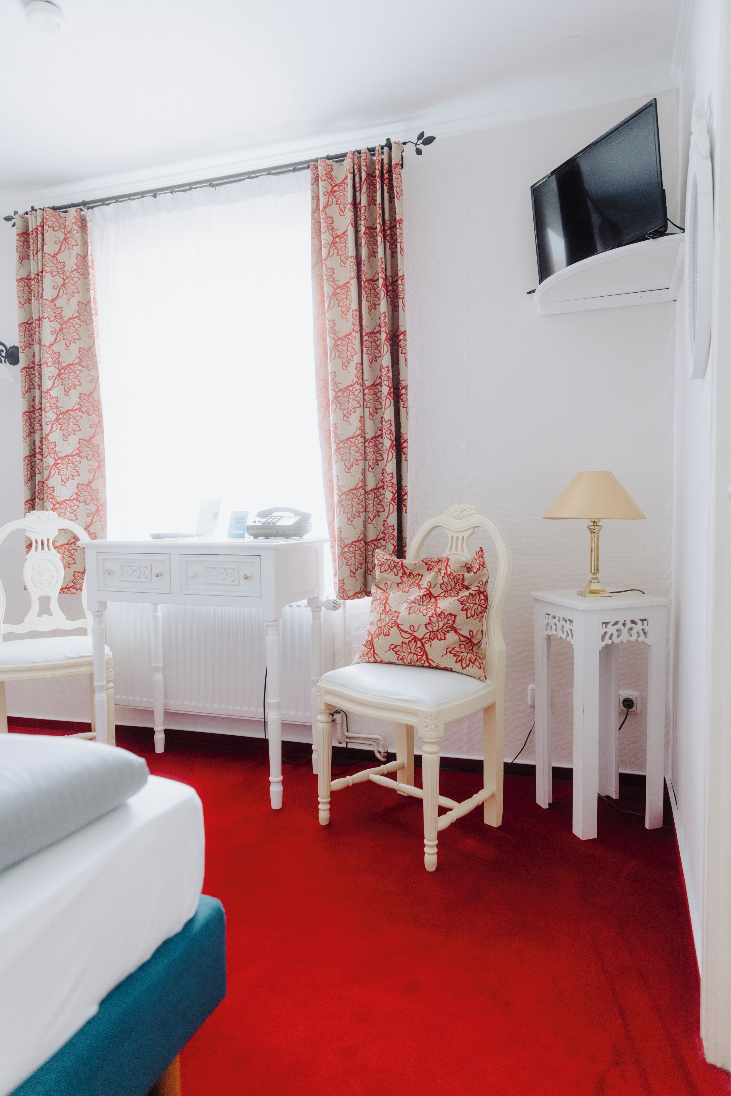 A hotel room with a bed, a desk with a chair, a television mounted on the wall, and a lamp on a side table.