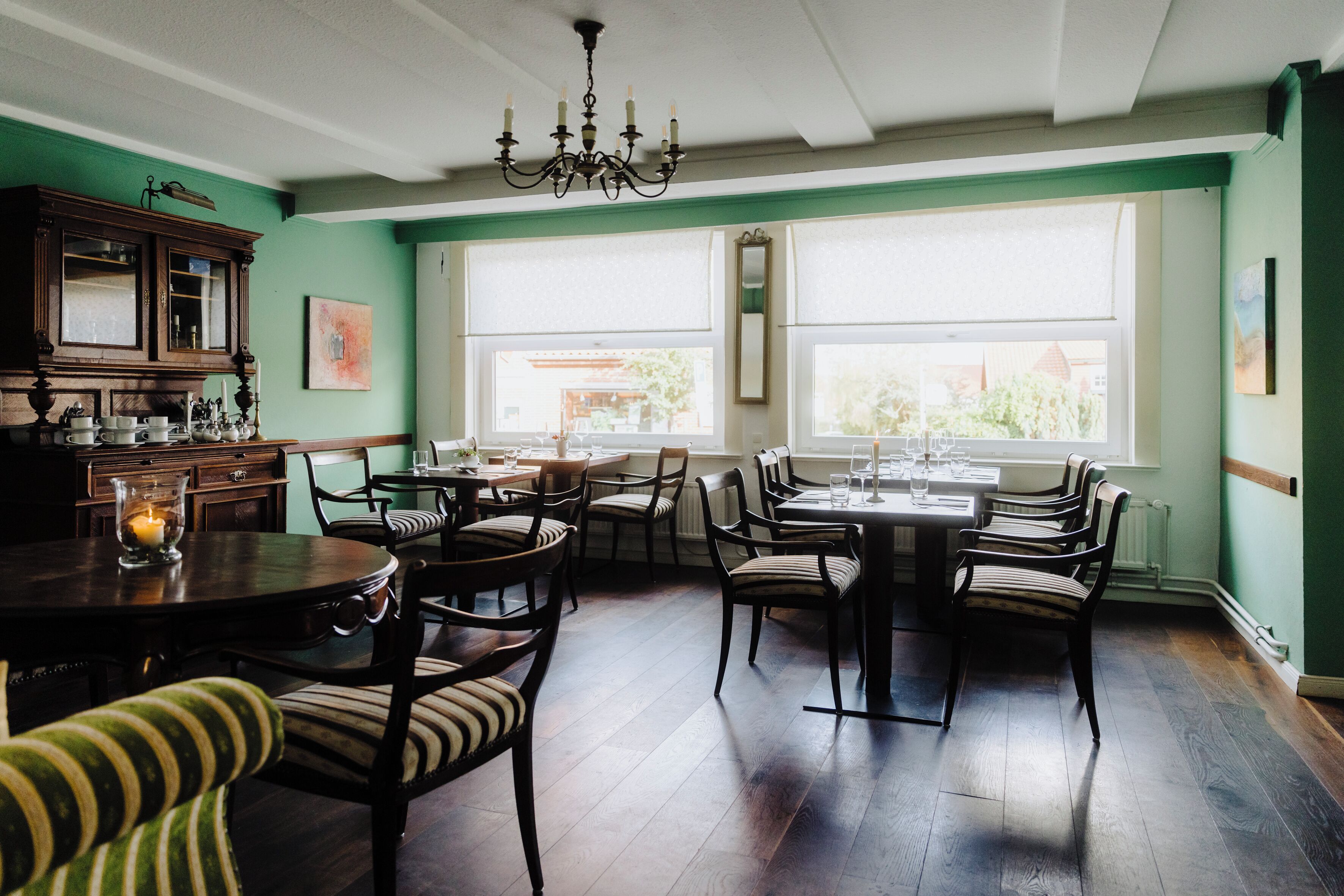 A dining area with a round table, chairs, and a chandelier. There are windows with blinds and a view of the outside.