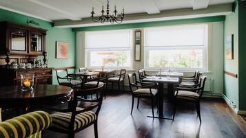 A dining area with a round table, chairs, and a chandelier. There are windows with blinds and a view of the outside.