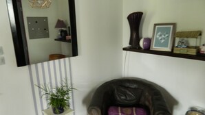 Un salon comprenant un fauteuil en cuir foncé, une petite table d’appoint blanche ornée d’une plante, un miroir au mur et une étagère avec des objets décoratifs.