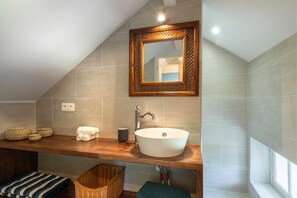 Un bagno con soffitto spiovente, un mobile lavabo in legno, un lavandino bianco, uno specchio con cornice di legno e una finestra.