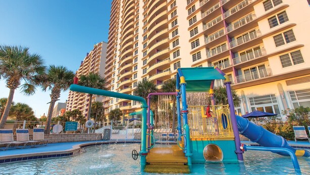 Ein Wasserpark mit Rutsche und Spielgeräten, umgeben von einem Pool und Palmen, mit einem hohen Gebäude im Hintergrund.