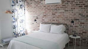 Ein Schlafzimmer mit Backsteinwand, einem Bett mit weißer Bettwäsche, einem Kleiderständer und einem Nachttisch mit einer Uhr.