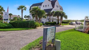 A large, two-story house with a white exterior and dark roof, surrounded by lush greenery and palm trees.