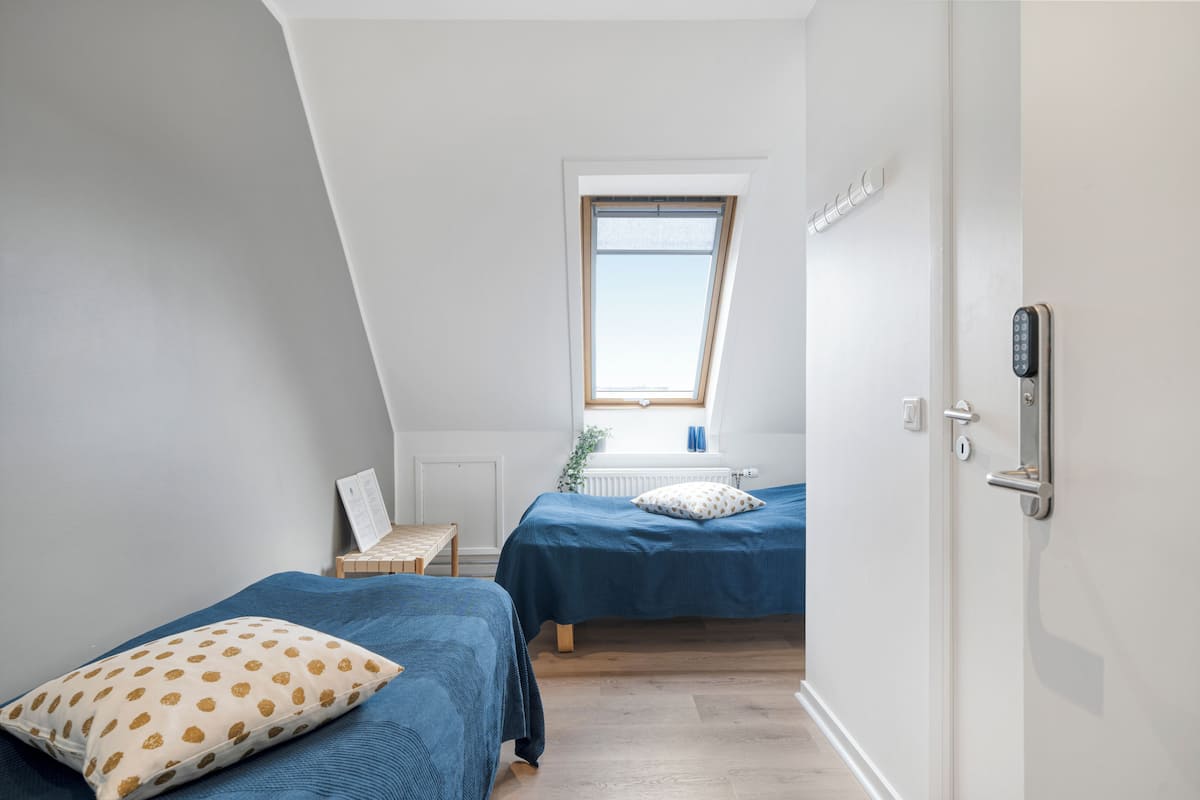 Un dormitorio pequeño y bien iluminado con dos camas, una ventana y una puerta con cerradura electrónica.