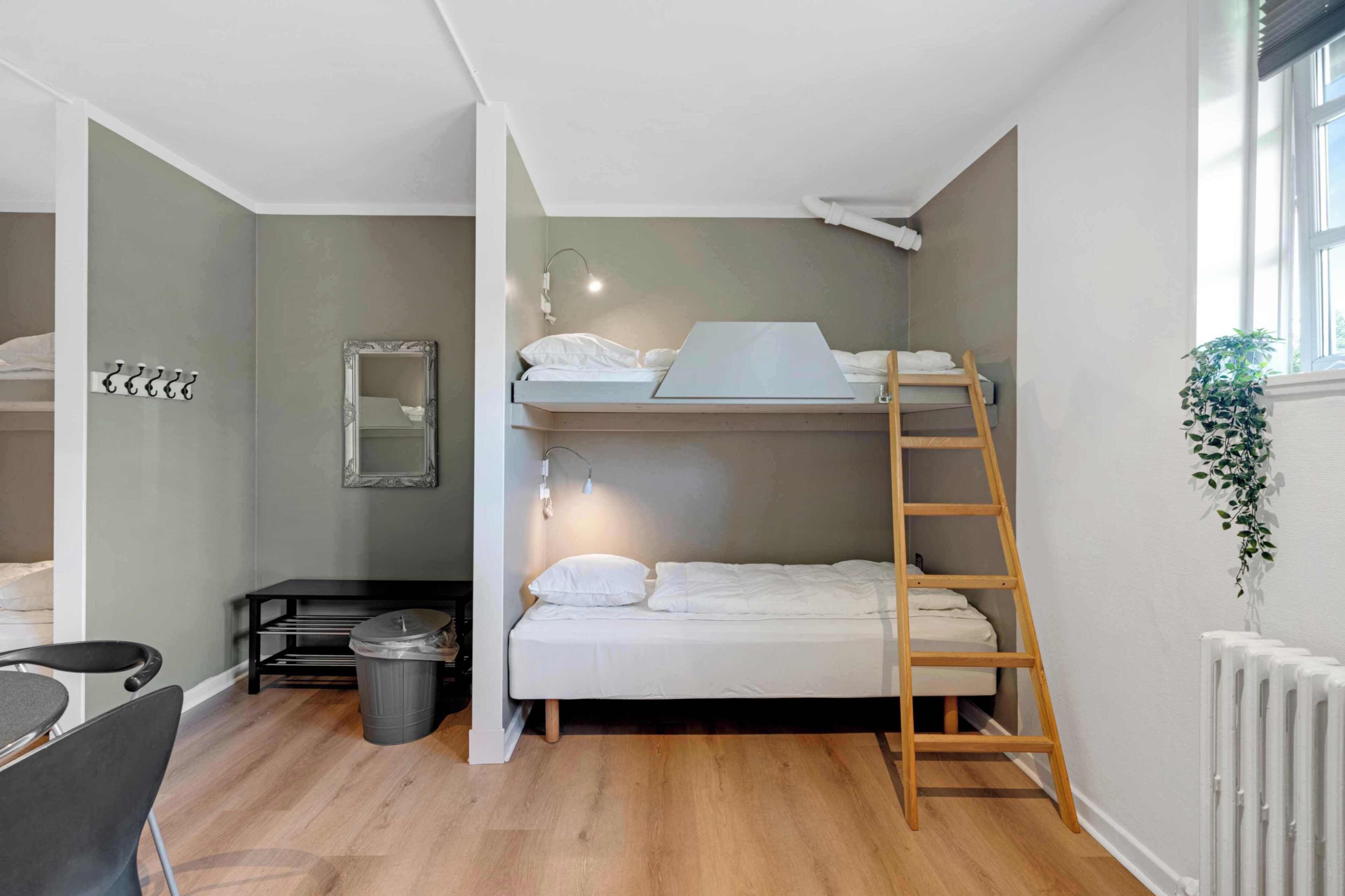 A bunk bed room with a wooden ladder, a desk with a chair, a trash bin, and a radiator.