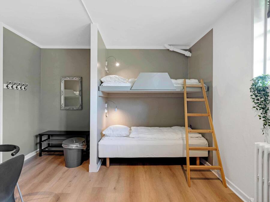 Habitación con literas, una escalera de madera, un escritorio con silla, un bote de basura y un radiador.