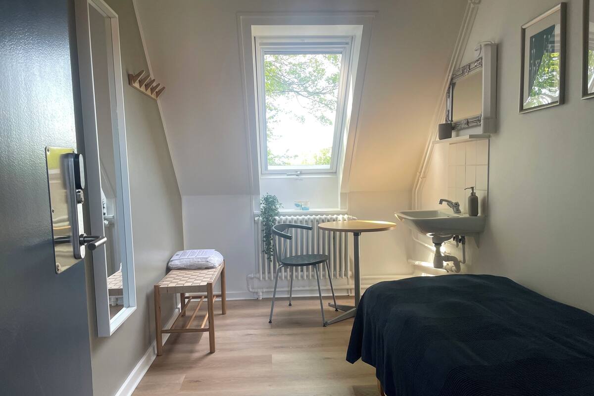 Una habitación pequeña con una cama, una mesita, una silla, un radiador y una ventana.