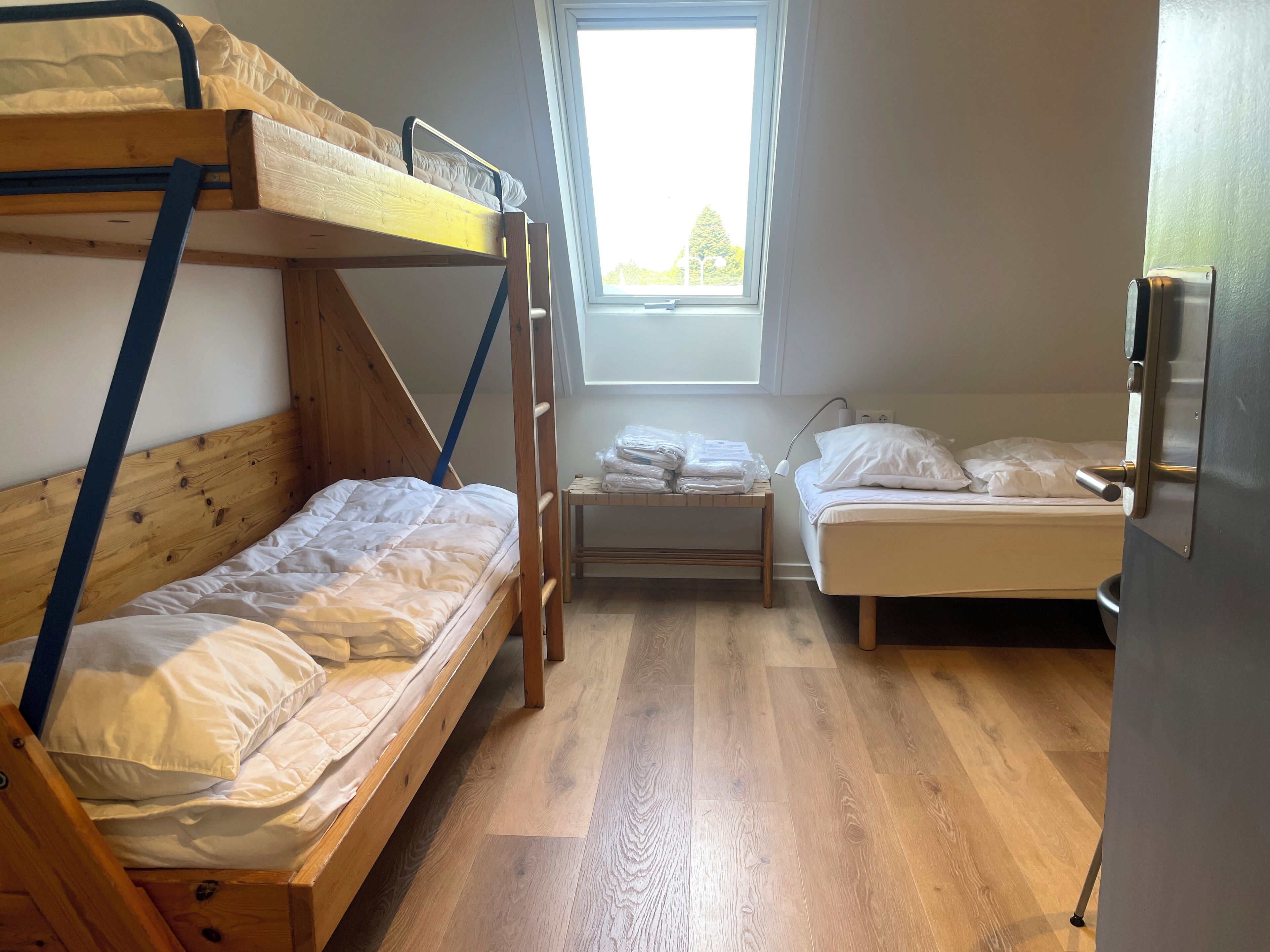 Une chambre de dortoir avec des lits superposés, une petite table sur laquelle sont pliées des serviettes et une fenêtre laissant entrer la lumière naturelle.