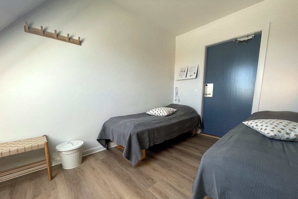 Una habitación con dos camas, un banco de madera, un cubo de basura blanco y una puerta azul.