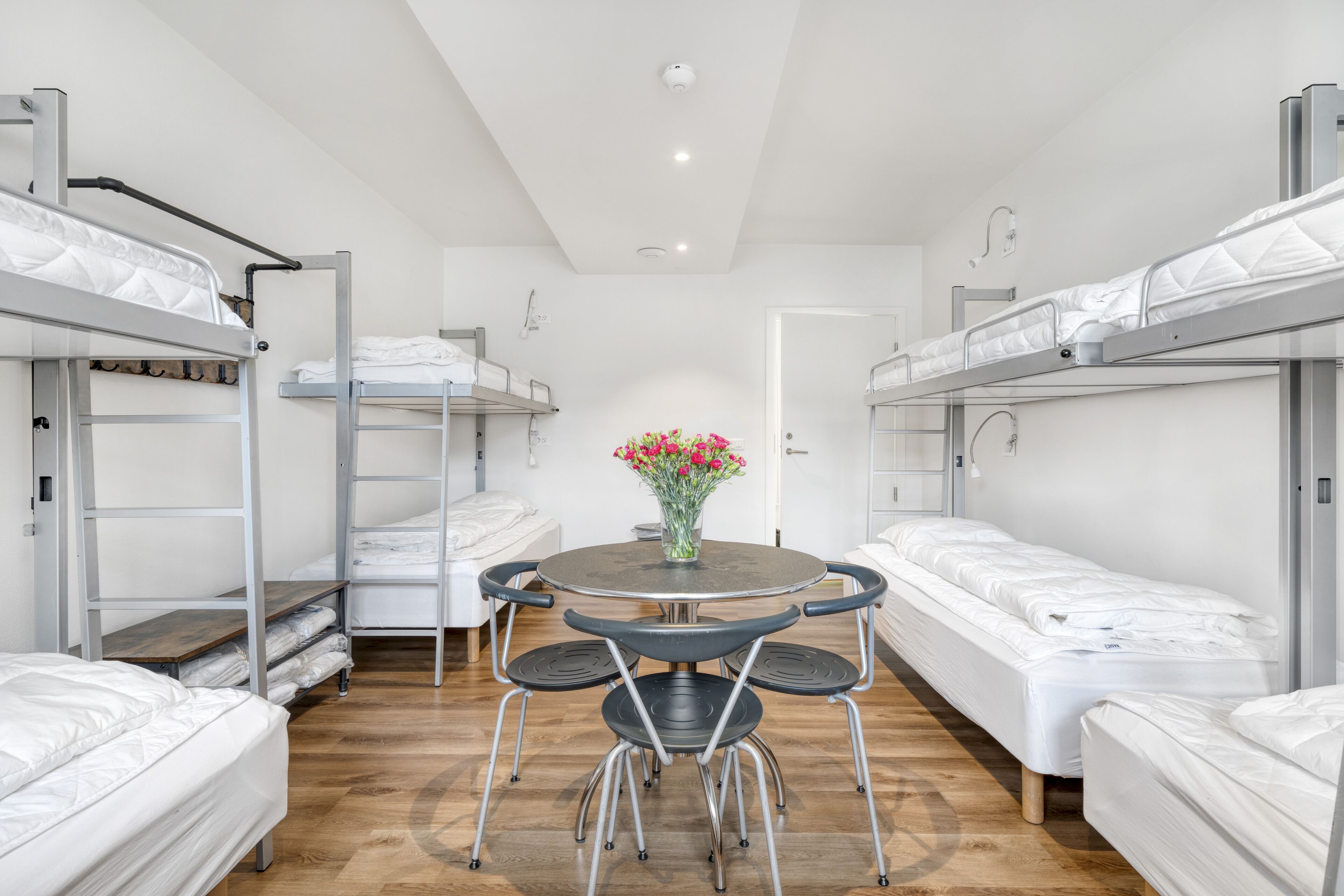 A dormitory room with bunk beds, a round table with chairs, and a vase with flowers.