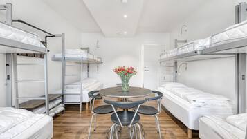 A dormitory room with bunk beds, a round table with chairs, and a vase with flowers.