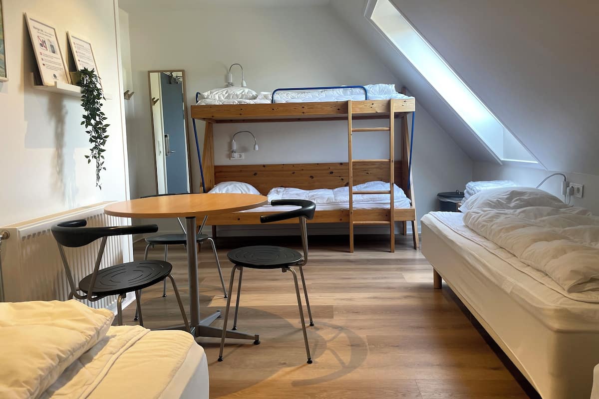 Una habitación tipo dormitorio con literas, una mesita pequeña y sillas, un radiador y un tragaluz.