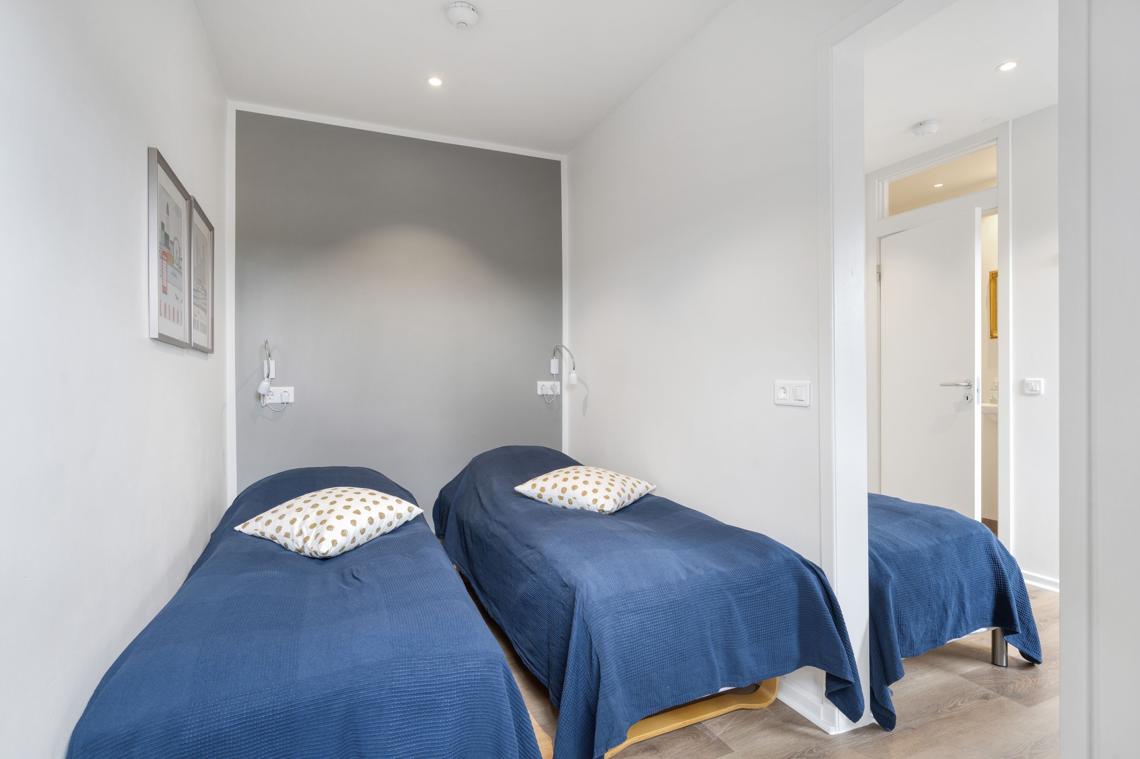 Una habitación con dos camas individuales, cada una con cubrecama azul y almohada blanca con lunares dorados. Las paredes son blancas y el suelo de madera.