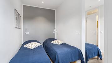 Una habitación con dos camas individuales, cada una con cubrecama azul y almohada blanca con lunares dorados. Las paredes son blancas y el suelo de madera.