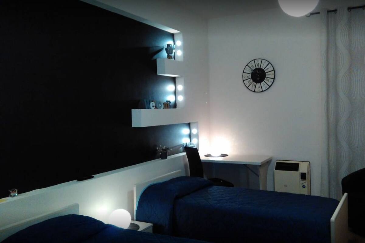 A bedroom with two beds, a desk with a computer, a wall clock, and a mirror with lights.