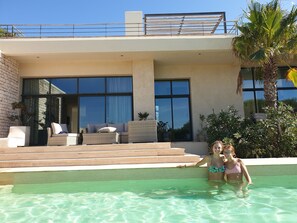 Un área moderna junto a la piscina con una piscina cristalina, mobiliario exterior y vistas a un edificio contemporáneo con amplias ventanas.