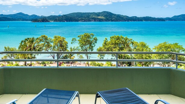 A balcony with two blue lounge chairs overlooking a turquoise sea and a forested island.