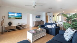A modern living room with a blue sofa, a coffee table, and a wooden cabinet. There is a ceiling fan, a painting of a beach, and a dining area in the background.
