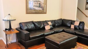 A black leather sectional sofa with a matching ottoman, a small wooden side table with a lamp, and two framed pictures on the wall. - ⭐Great4Stargazing ⭐ Steps2Lake ⭐ Perfect4Families ⭐Patio (Kingsland)