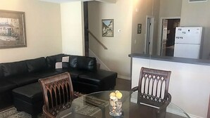 A living room with a black leather sofa, a glass coffee table with decorative items, and a dining area with two wooden chairs. A kitchen area with a refrigerator is visible in the background. - ⭐Great4Stargazing ⭐ Steps2Lake ⭐ Perfect4Families ⭐Patio (Kingsland)