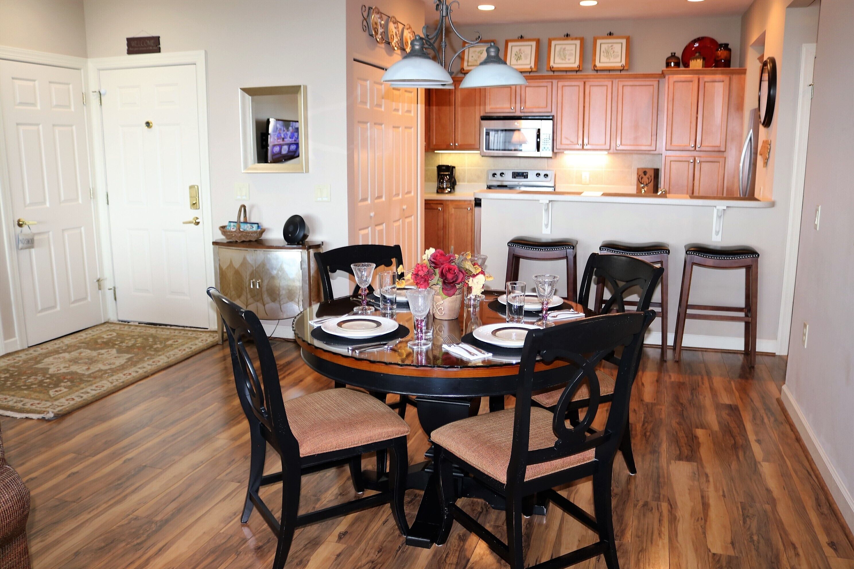 A dining area with a round table set for four, surrounded by wooden chairs. A kitchen with wooden cabinets and a microwave is visible in the background.