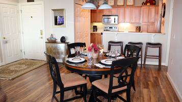 A dining area with a round table set for four, surrounded by wooden chairs. A kitchen with wooden cabinets and a microwave is visible in the background.