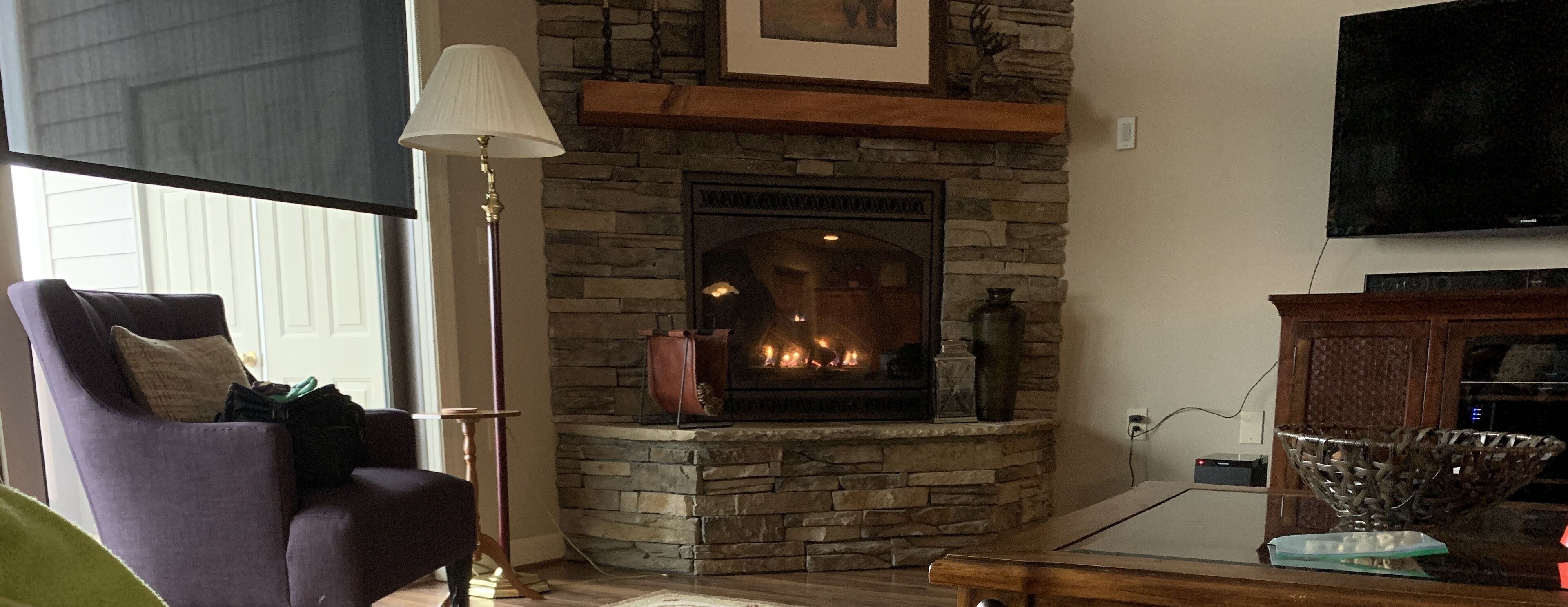 A living room with a stone fireplace, a television mounted above it, a floor lamp, and a sofa with a cushion.