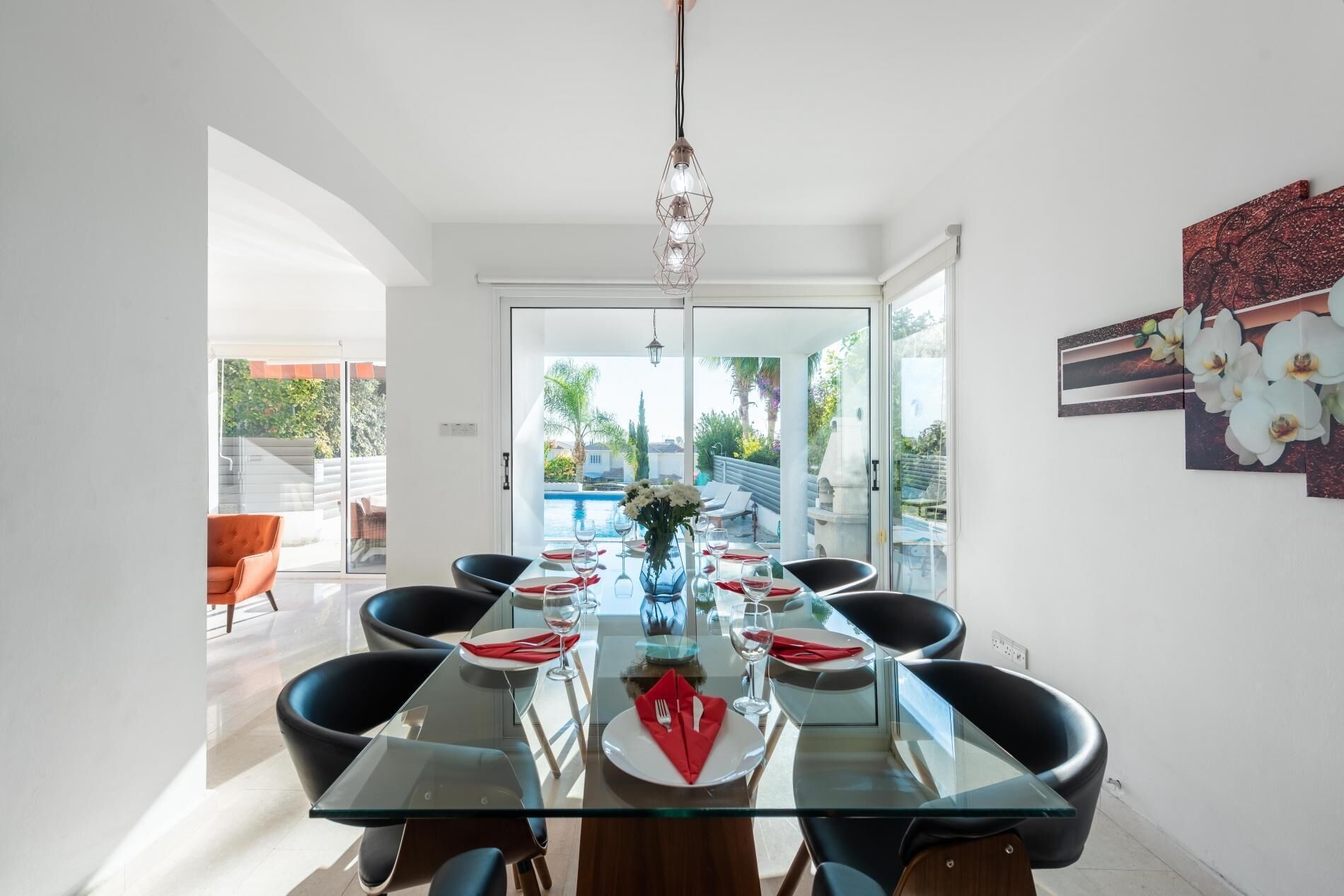 A dining area with a glass table, black chairs, and a view of a swimming pool and greenery outside.