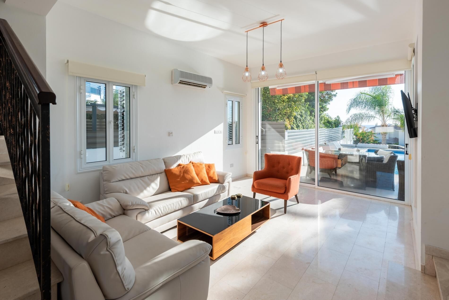 A spacious living room with a sofa, armchair, and coffee table. There is a sliding glass door leading to an outdoor area with a pool.