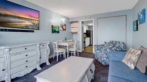 Un salon avec une commode blanche, une table à manger avec des chaises, un canapé bleu orné de coussins à motifs floraux et un grand tableau représentant un coucher de soleil sur une jetée.
