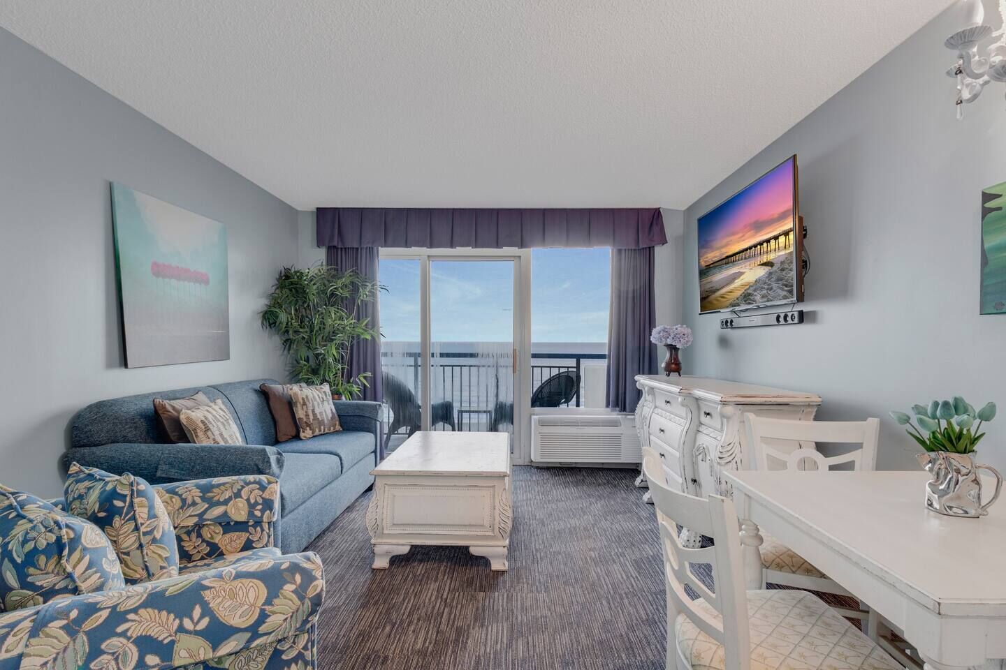 A living room with a sofa, coffee table, and a dining area with a table and chairs. There is a balcony with a view of the sea.