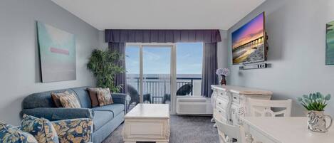 A living room with a sofa, coffee table, and a dining area with a table and chairs. There is a balcony with a view of the sea.