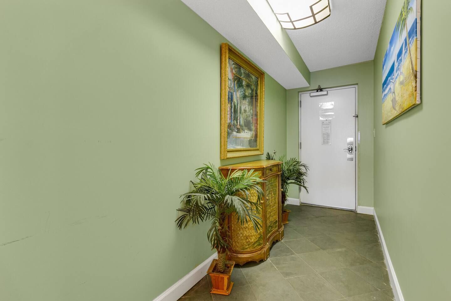 Un couloir avec une console en bois, deux plantes en pot, un tableau encadré et une porte blanche.