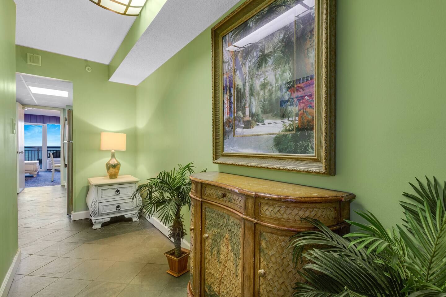 A hallway with a green wall, a wooden cabinet, a white dresser, a lamp, and a large framed picture.