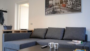 A modern living room with a grey sectional sofa, a coffee table with remote control, mugs, and a magazine. A black and white cityscape photo is on the wall.