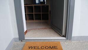 A doorway with a welcome mat and a tiled floor leading to a room with a wooden shelf and a basket.
