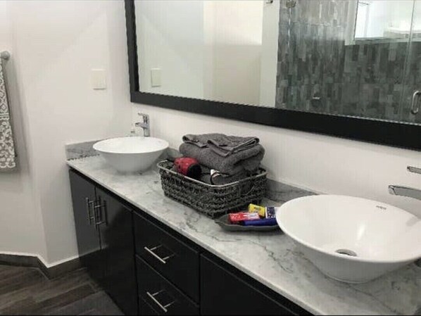 Un baño con dos lavamanos, un espejo grande y una cesta con toallas y artículos de aseo.