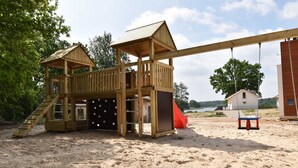 A wooden play structure with a slide and climbing areas, set on sandy ground with trees and a house in the background.