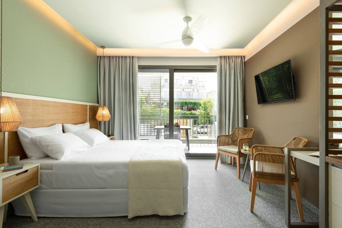 A hotel room with a bed, bedside lamps, a TV, a balcony with chairs, and a view of the outside.